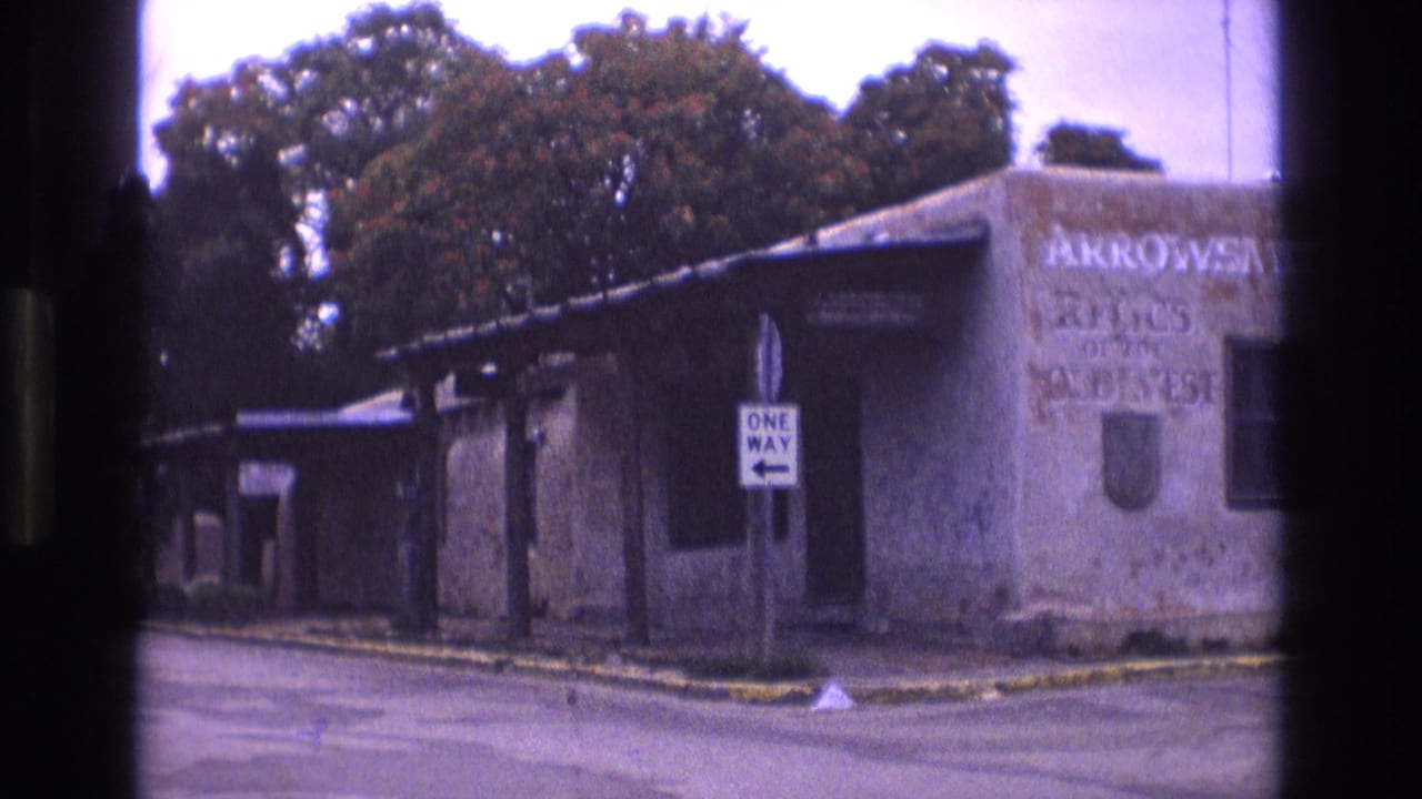 1973 New Mexico 8mm frame with a weathered roadside building, readable Arrowsmith relics lettering, a one-way sign, and trees above the roofline.