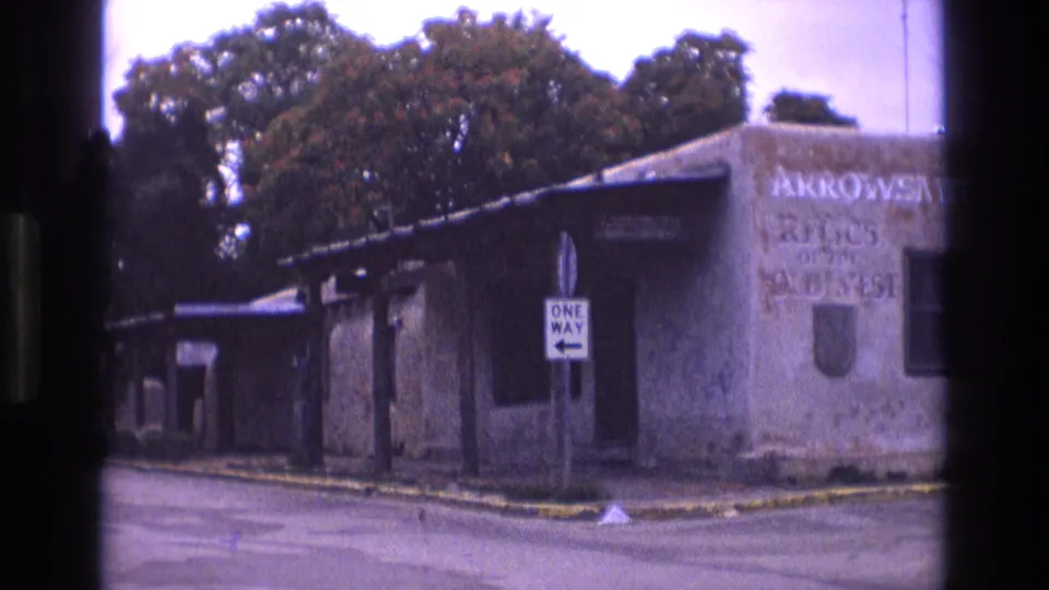 1973 New Mexico 8mm frame with a weathered roadside building, readable Arrowsmith relics lettering, a one-way sign, and trees above the roofline.