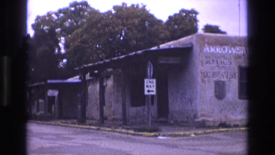 1973 New Mexico 8mm frame showing a closer roadside view of the weathered building, white one-way sign, covered walkway, and faded Old West relics lettering.