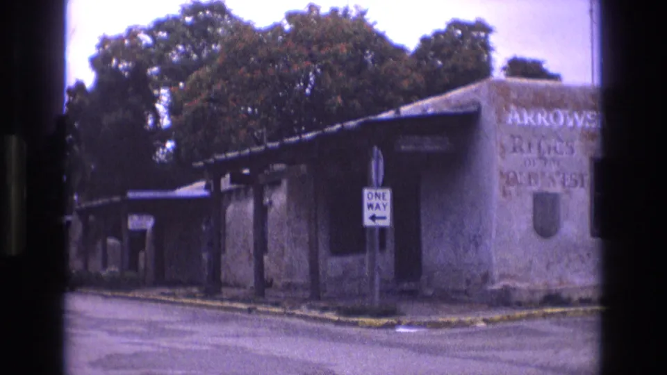 1973 New Mexico 8mm frame showing the street corner from a slightly shifted angle, with the one-way sign centered near the weathered adobe-style storefront.