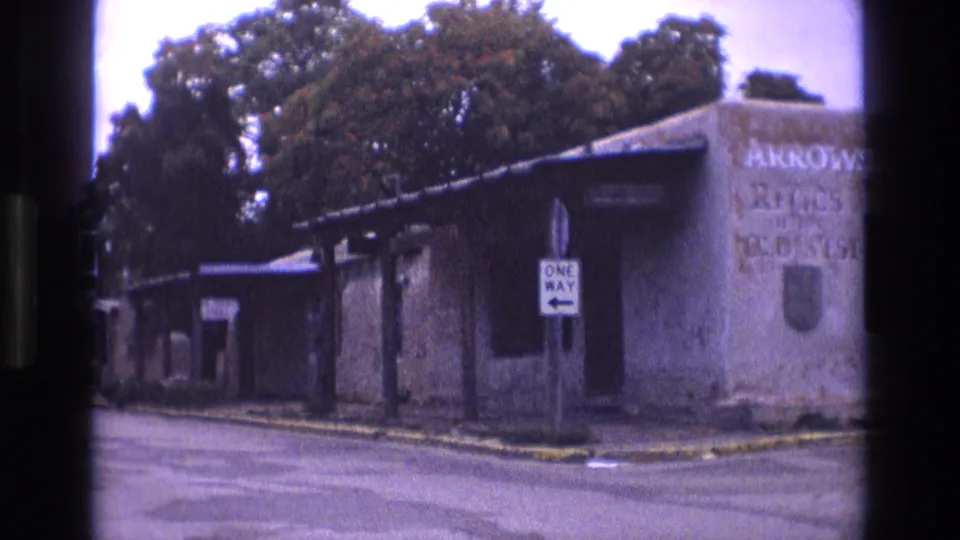 1973 New Mexico 8mm frame showing a weathered adobe corner building, street pavement, a one-way sign, and partially readable painted landmark text.