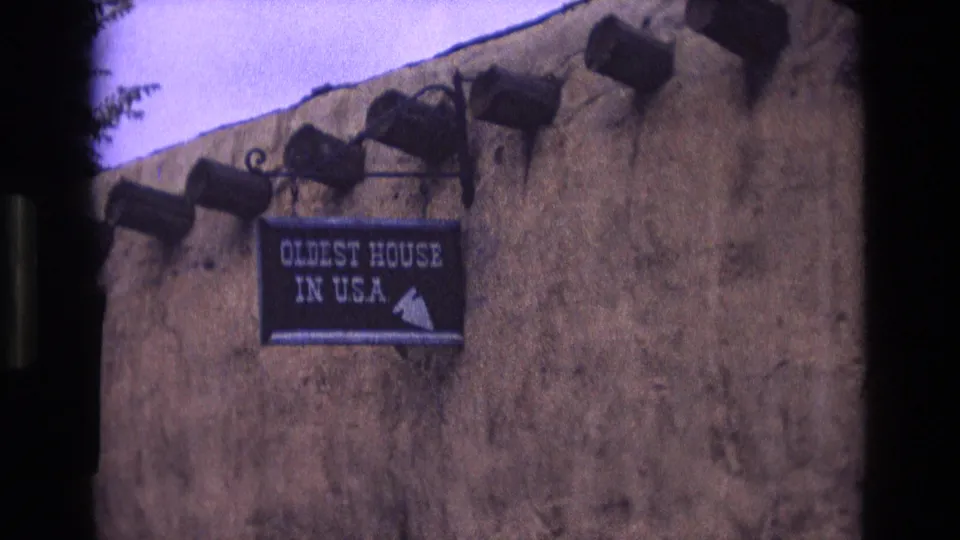 1973 New Mexico 8mm frame showing a closer visitor-shot view of the "OLDEST HOUSE IN USA" sign against a weathered adobe wall with exposed wooden beams.
