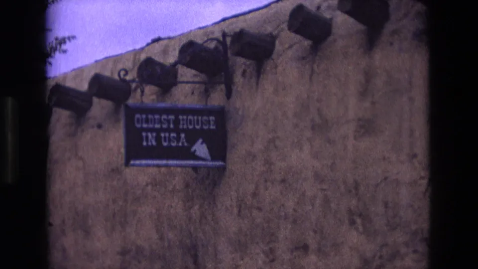 1973 New Mexico 8mm frame showing a close visitor view of an adobe exterior wall with projecting beams and a sign reading "Oldest House in USA."