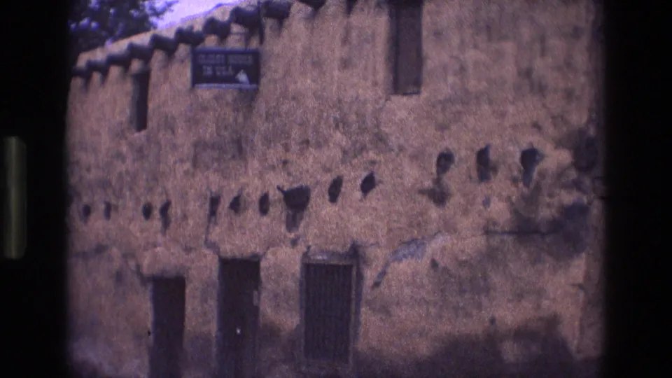 1973 New Mexico home-movie frame showing a wider exterior of the adobe building, with the readable sign near the upper wall and dark film borders from the scan.