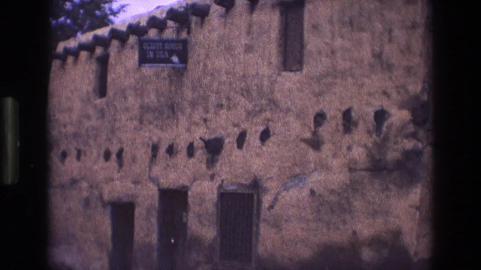 1973 New Mexico 8mm frame showing a weathered adobe building facade with small wall openings, dark doorway forms, a barred window, and a sign reading "OLDEST HOUSE IN USA."