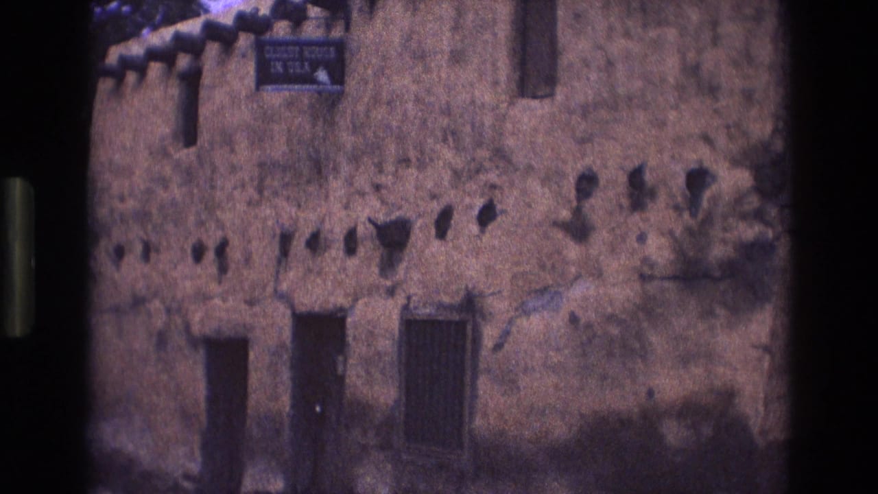 Adobe wall exterior with sign near the roofline and dark door and window openings below.