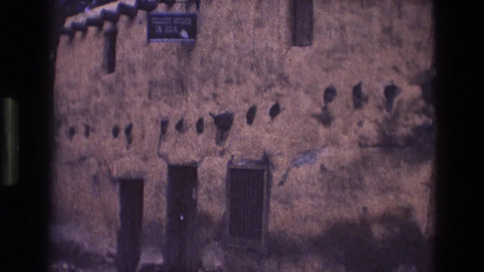 1973 New Mexico 8mm frame showing the lower portion of a weathered adobe building, including a readable historic-attraction sign, a dark doorway, and a barred window.