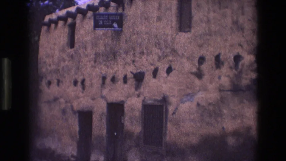 1973 New Mexico 8mm frame of a historic adobe exterior with the "OLDEST HOUSE IN USA" sign, weathered stucco surface, small openings, and strong shadow across the lower wall.