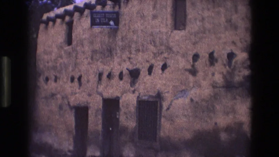 1973 New Mexico 8mm frame showing a weathered adobe building facade with rough plaster, small wall openings, dark wooden doors, and a sign reading "OLDEST HOUSE IN USA."