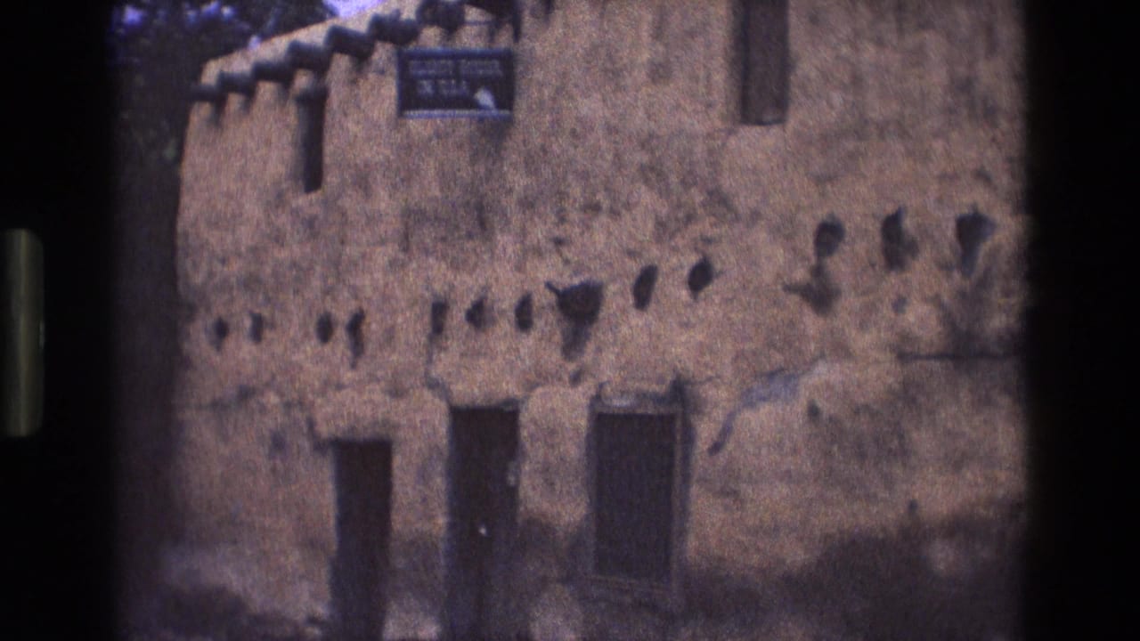 Similar view of the adobe building facade with doors, wall openings, and upper sign.