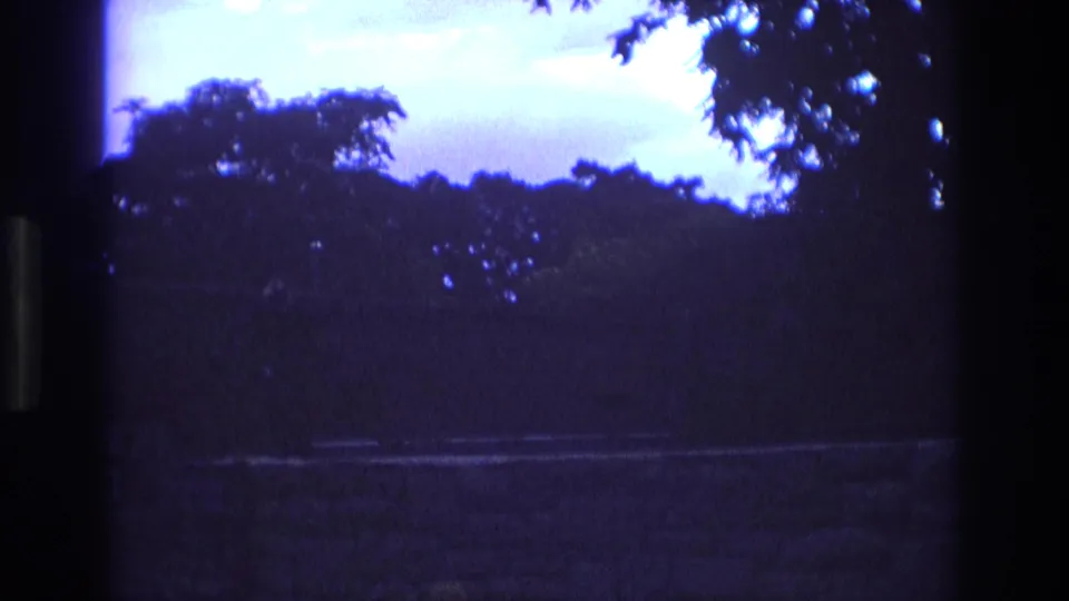 1973 New Mexico 8mm frame showing dark tree silhouettes, a blue-purple evening sky, and a faint horizontal path or sidewalk seen through a dark foreground edge.