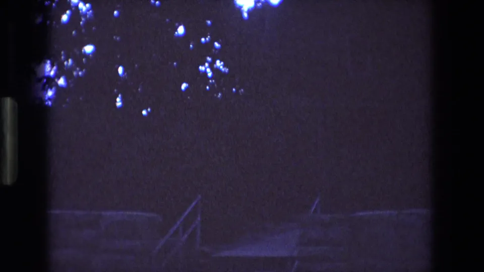 1973 New Mexico 8mm frame showing a very dark exterior view with a faint railing or stair structure, shadowed trees, and blue-white flare spots near the upper edge.