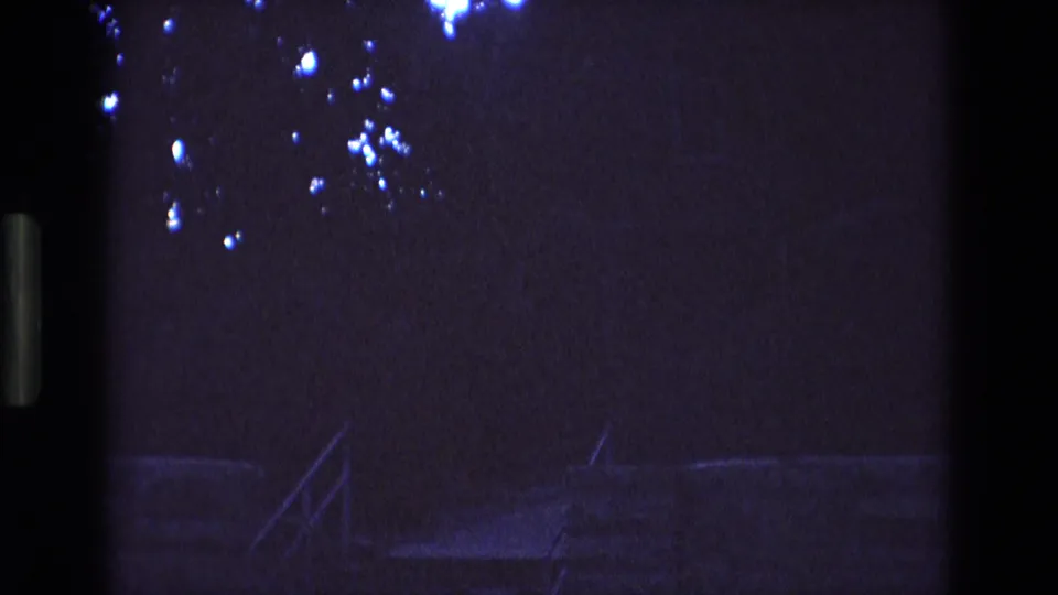 1973 New Mexico 8mm frame with a shadowed outdoor stair or railing form, scattered bright flare spots above, and a dark vertical edge dividing the view.