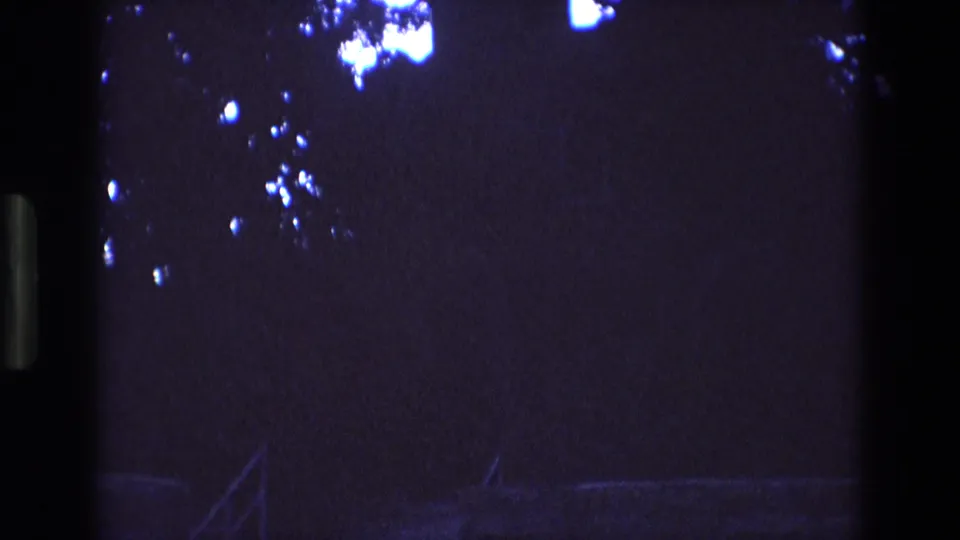 1973 New Mexico 8mm frame showing a nearly black exterior with faint diagonal railing lines, indistinct foliage, blue-white flare clusters, and visible film grain.