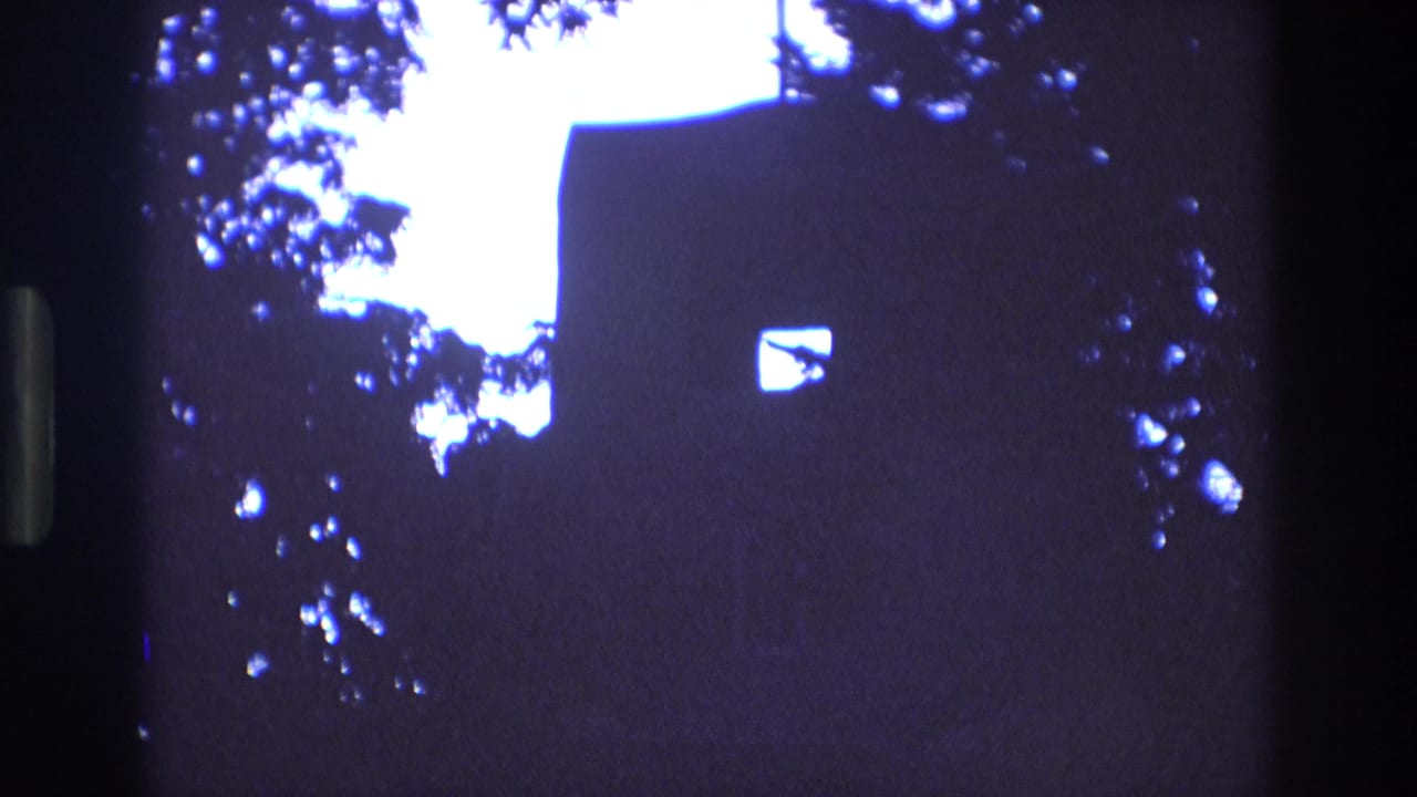 1973 New Mexico 8mm frame showing a dark silhouetted church wall with a small bright window, a roofline cross, and leafy branches against a pale sky.
