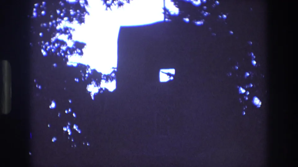 1973 New Mexico 8mm frame showing a dark silhouetted church wall with a small bright window, a roofline cross, and leafy branches against a pale sky.