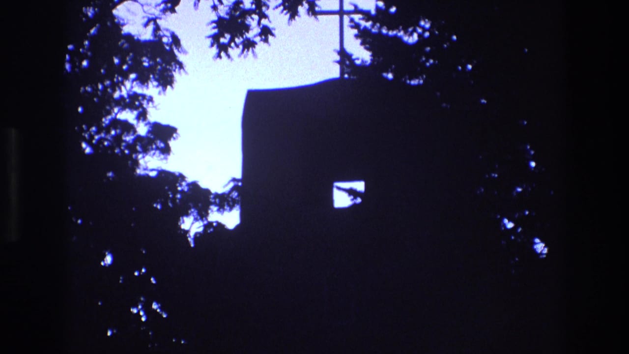 1973 New Mexico 8mm frame with a silhouetted church exterior, small rectangular window, cross near the roofline, and tree branches cutting into the bright sky.