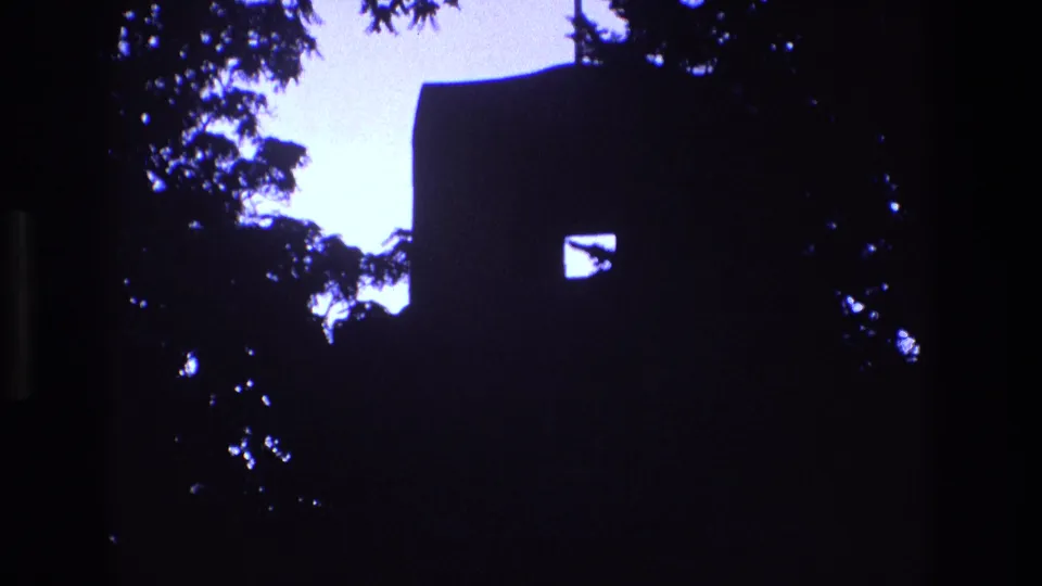 1973 New Mexico home-movie frame showing the church as a near-black silhouette with a small lit window shape, a cross above the roof, and foliage around the edges.