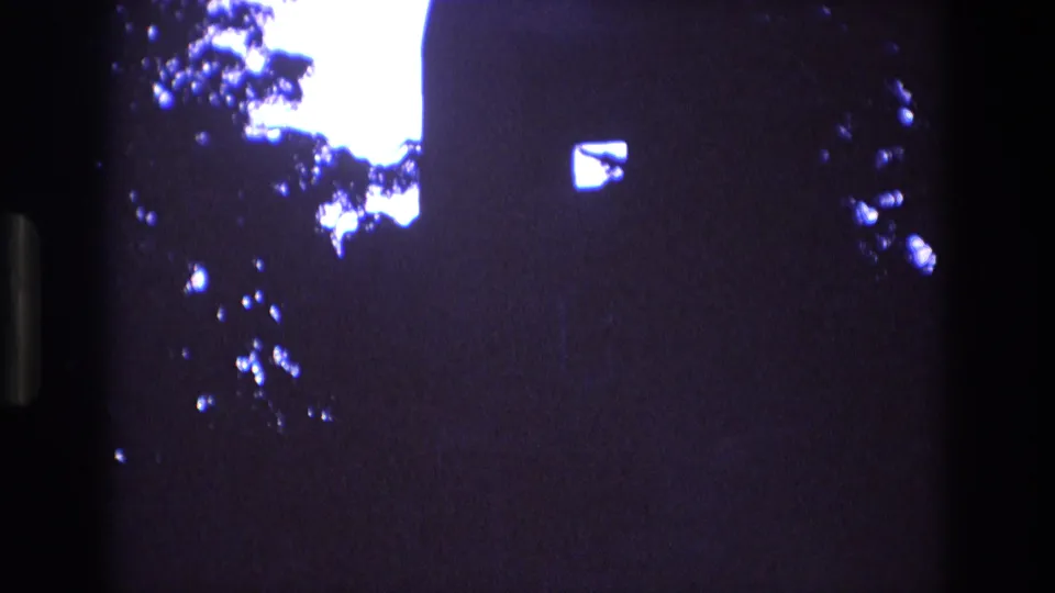 1973 New Mexico 8mm frame with blue-purple tint, a silhouetted church wall, small window opening, cross at the roofline, and leafy branches against the sky.