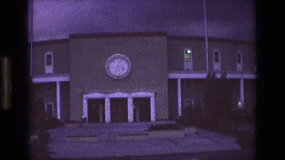 1973 New Mexico 8mm frame showing a large institutional-looking building at night with a circular emblem above the entrance, concrete walkways, stairs, railings, doors, and small lit windows.
