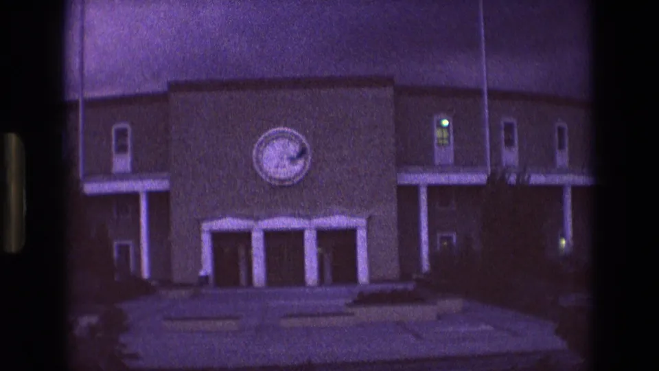 1973 New Mexico 8mm frame holding on the large building exterior at night, with the circular emblem, entrance doors, concrete steps and paths, railings, and faint window lights visible through the grain.