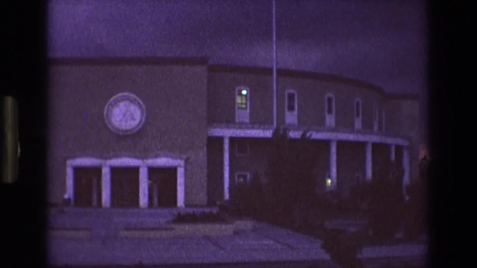 1973 New Mexico 8mm frame showing a large two-story building at twilight, with a curved facade, white porch pillars, illuminated windows, and a circular emblem-like feature above the entrance.
