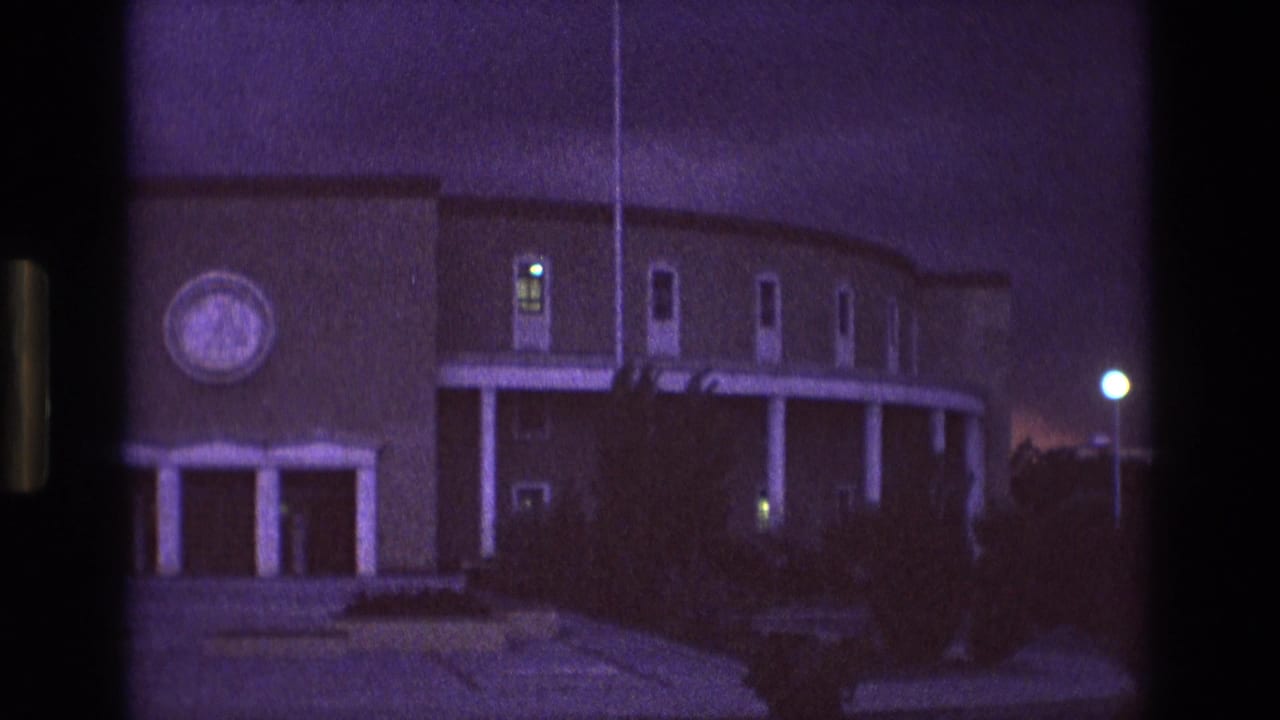 1973 New Mexico home-movie frame showing the building from a similar angle, with the circular feature partly visible, white pillars, glowing windows, shadowed shrubs, and a streetlight at right.