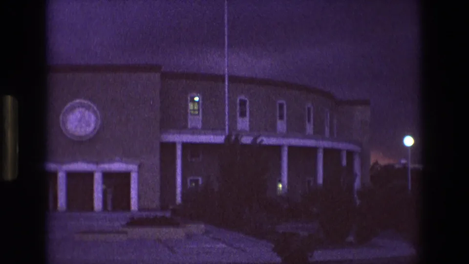 1973 New Mexico home-movie frame showing the building from a similar angle, with the circular feature partly visible, white pillars, glowing windows, shadowed shrubs, and a streetlight at right.