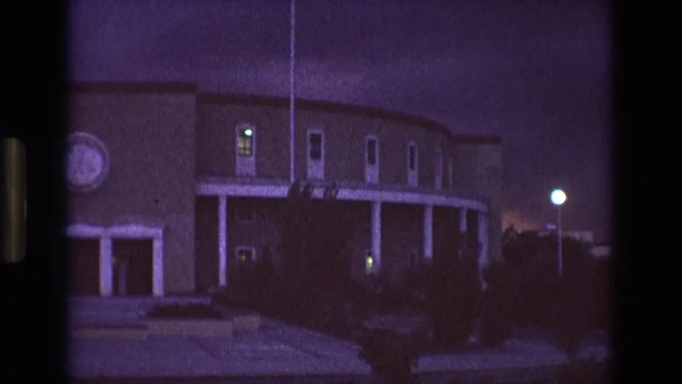 1973 New Mexico 8mm frame with the left side of the building partly cropped, showing the covered porch columns, several narrow windows, dark landscaping, and a bright streetlight against the dusk sky.
