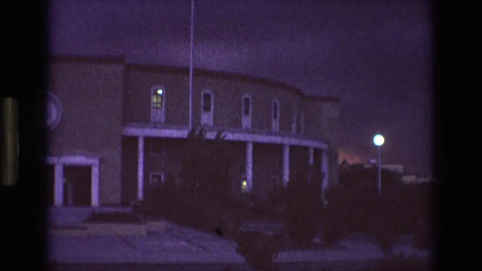 Final 1973 New Mexico 8mm frame of the twilight exterior, emphasizing the curved roofline, white porch columns, glowing windows, dark shrubs, and soft purple film color.