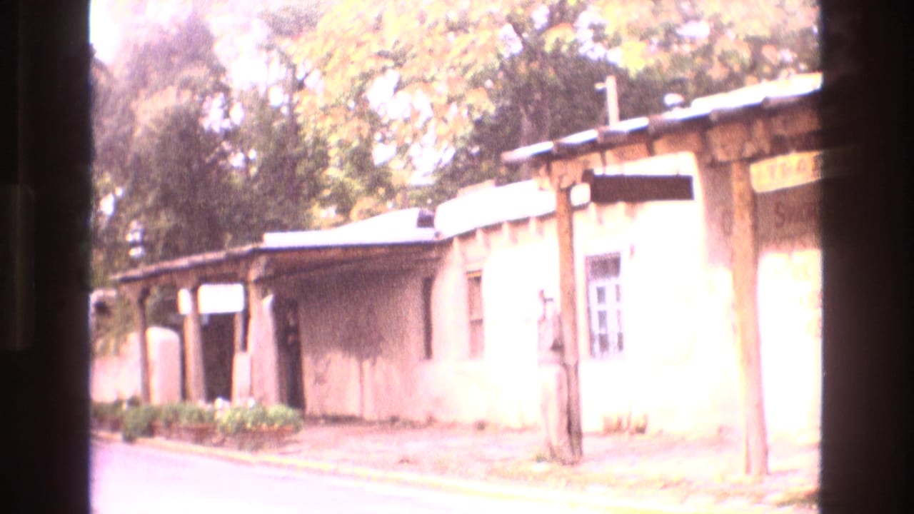 1973 New Mexico home-movie frame showing an adobe-style building with a wooden porch, street curb, landscaping, and trees with autumn foliage.