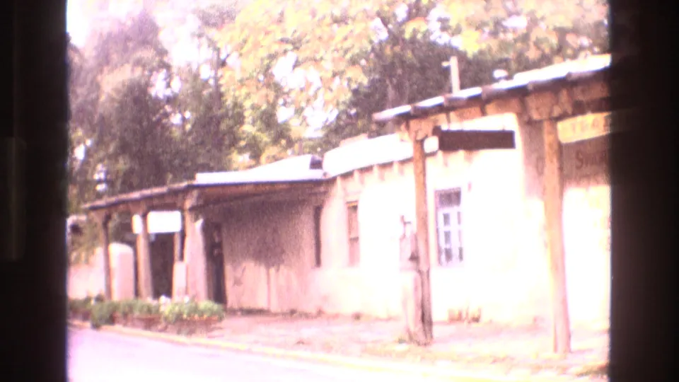 1973 New Mexico home-movie frame showing an adobe-style building with a wooden porch, street curb, landscaping, and trees with autumn foliage.