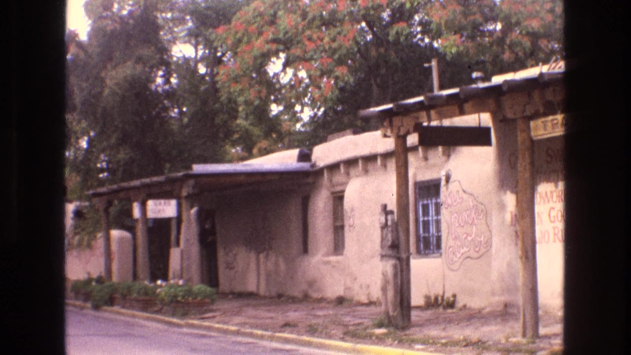 1973 New Mexico frame of an adobe-style storefront or residence with porch posts, a barred window, hand-painted signage, and leafy trees in the background.