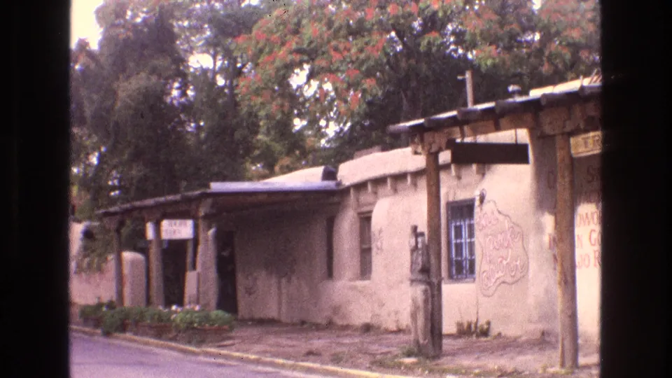 1973 New Mexico home-movie frame showing a closer street view of an adobe-style building with a wooden porch, blue-framed barred window, painted wall sign, and low plantings.