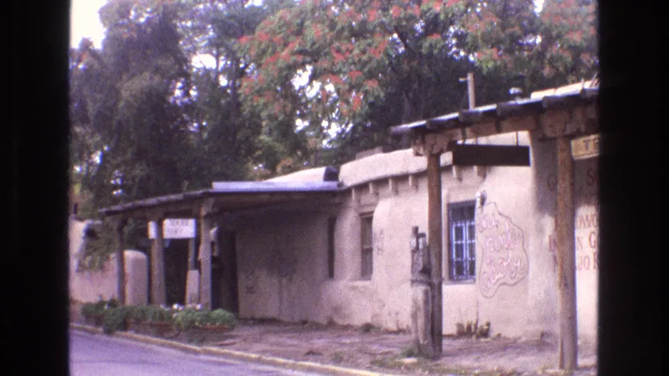 1973 New Mexico frame showing the adobe-style exterior from across the street, including porch posts, a barred window, hand-painted wall signage, curb, and autumn foliage behind the roofline.