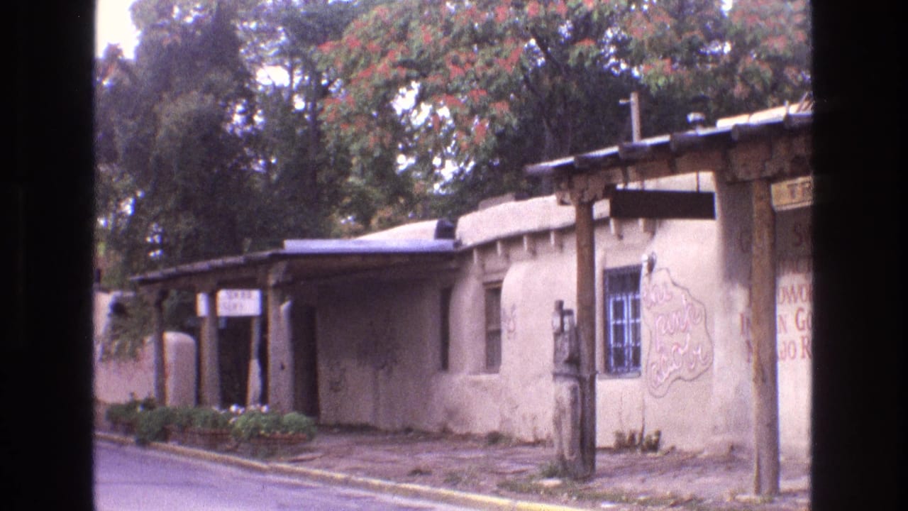 1973 New Mexico 8mm frame showing a tan stucco adobe-style roadside building with a covered porch, trees with red blossoms, and part of a hand-painted 'The Pink Adobe' sign.