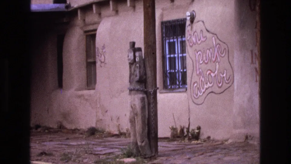 Closer 1973 New Mexico home-movie frame showing the adobe-style wall, a barred window, a carved wooden figure near the entrance area, and the hand-painted 'The Pink Adobe' sign.