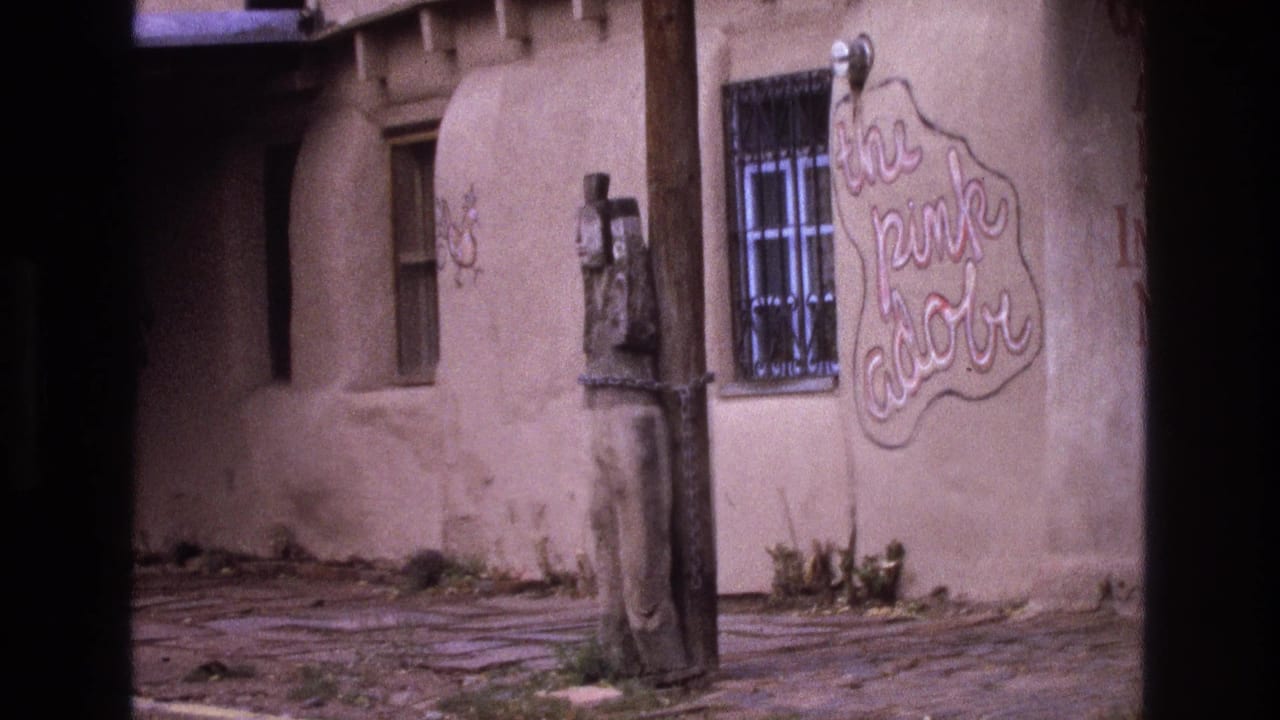 1973 New Mexico 8mm frame showing an adobe exterior with a barred window, weathered wooden pole, chained carved figure, and cursive sign reading "the pink adobe."