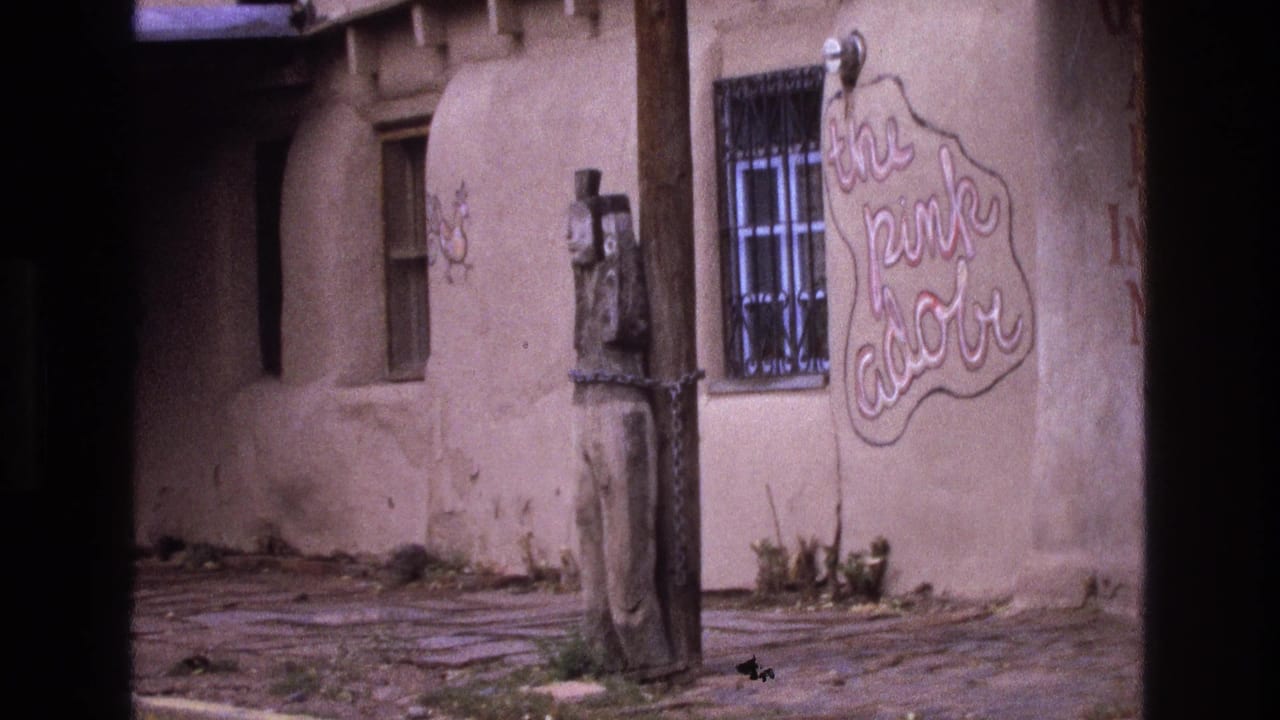 1973 New Mexico home-movie frame of a rustic adobe building exterior with a barred window, a wooden pole, a chained carved figure, and a cursive "the pink adobe" sign.