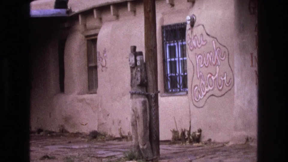 1973 New Mexico 8mm view of the same adobe exterior, with soft archival color, a barred window, wood pole, chained carved figure, and hand-painted sign.