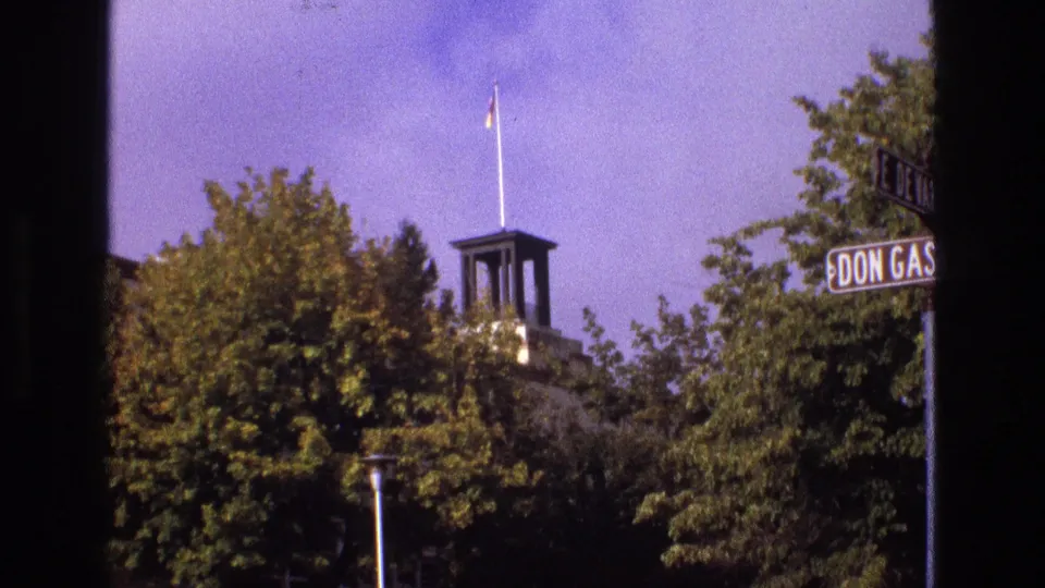 1973 New Mexico 8mm frame showing trees, a small tower or cupola, a flagpole, and a cropped street or place sign near the edge of the frame.