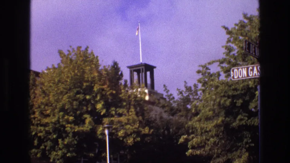 1973 New Mexico 8mm frame showing a small tower or monument with a flagpole rising above leafy trees, with partial street signs at the right edge.