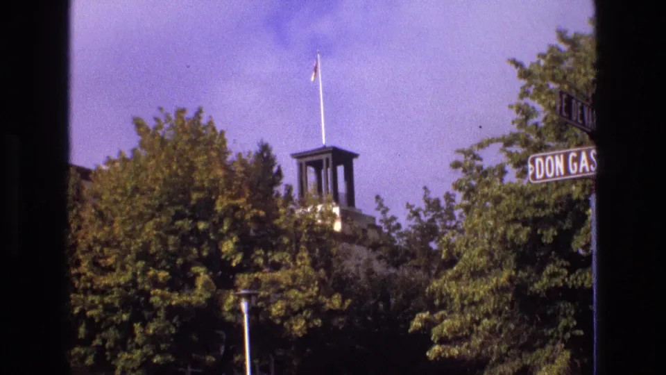 1973 New Mexico 8mm frame looking past trees toward a small civic-looking tower with a flagpole, a pole in the foreground, and partial signage at the right.