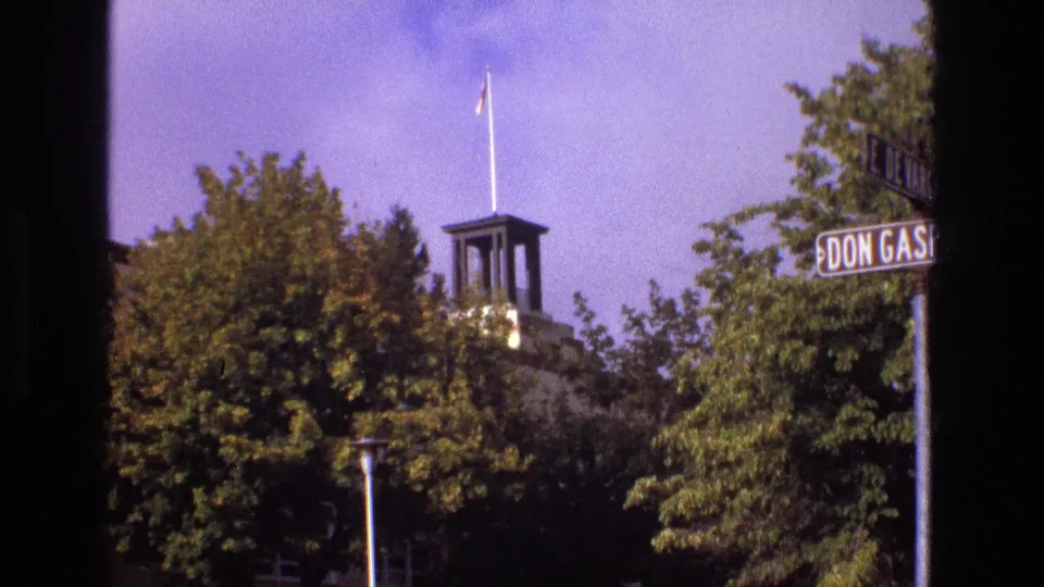 1973 New Mexico home-movie frame showing a small flag on a pole above a tower or monument, partly hidden by trees and bordered by a cropped street sign.