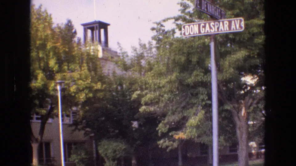 1973 New Mexico 8mm frame looking toward the Don Gaspar Avenue sign, dense roadside trees, and a towered structure partly hidden by foliage and film-edge shadow.