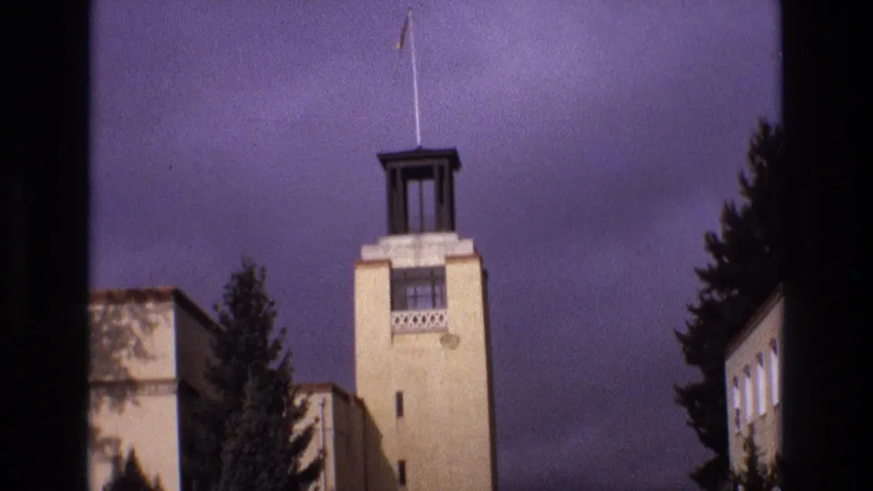 1973 New Mexico 8mm frame with a low-angle view of a pale tower or steeple, nearby building edges, trees, and a dark clouded sky.