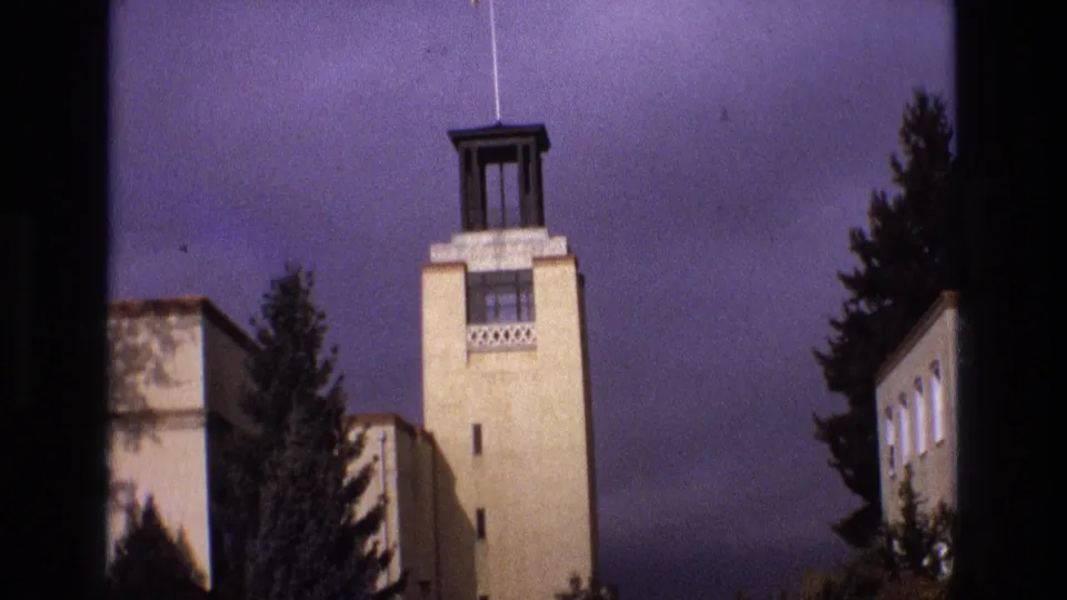 1973 New Mexico 8mm frame showing a tall beige institutional-looking building with a dark rooftop tower element, decorative lattice detail, and evergreen trees in the foreground.