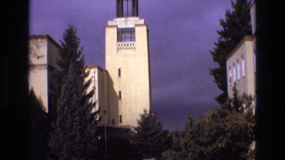1973 New Mexico 8mm frame showing the upper portion of the beige building tower, decorative window area, evergreen trees, and foreground shrubs with soft home-movie grain.