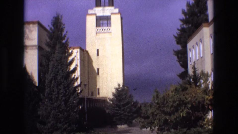 1973 New Mexico 8mm frame showing the beige tower and adjacent institutional-looking walls with evergreen trees and shrubs filling the lower edge of the composition.