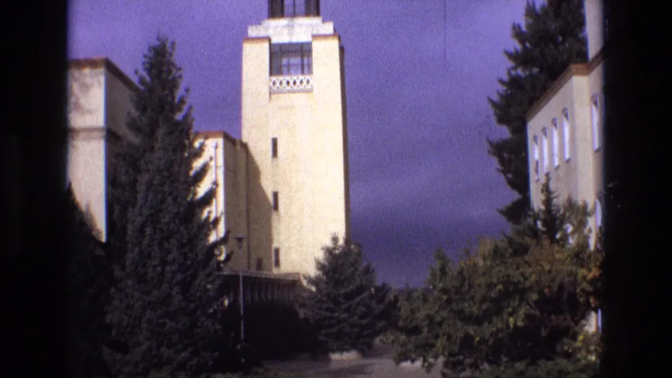 1973 New Mexico 8mm frame showing a pale towered building with geometric detailing, lower wings, evergreen trees, and a color-shifted sky.